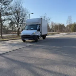 Biały samochód dostawczy Iveco z zabudową kontenerową zaparkowany na parkingu w słoneczny dzień, widoczne drzewa w tle.