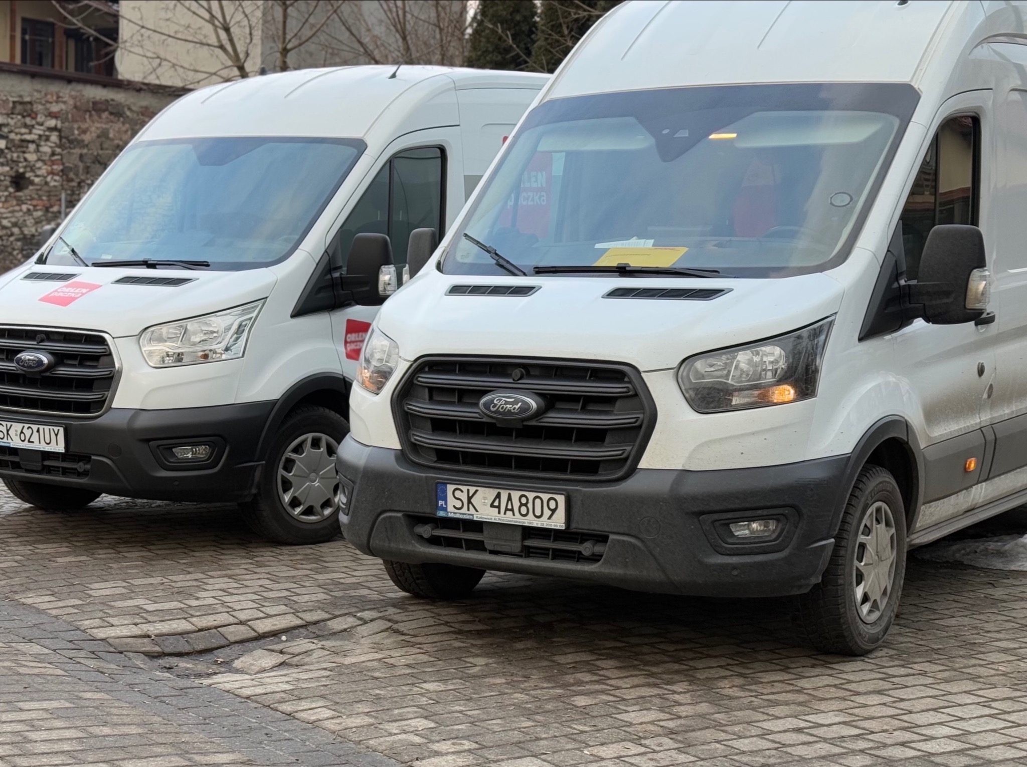 Dwa białe busy Ford Transit zaparkowane na brukowanej ulicy, jeden z logo Orlen Paczka na boku, drugi z żółtą kartką za szybą, widoczne numery rejestracyjne.
