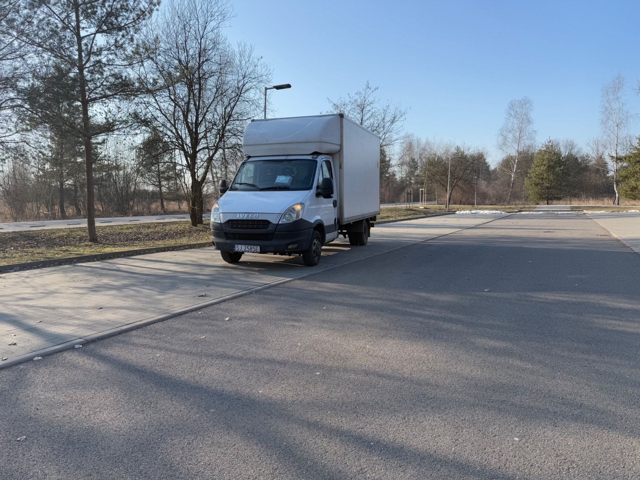 Biały samochód dostawczy Iveco z zabudową kontenerową zaparkowany na parkingu w słoneczny dzień, widoczne drzewa w tle.