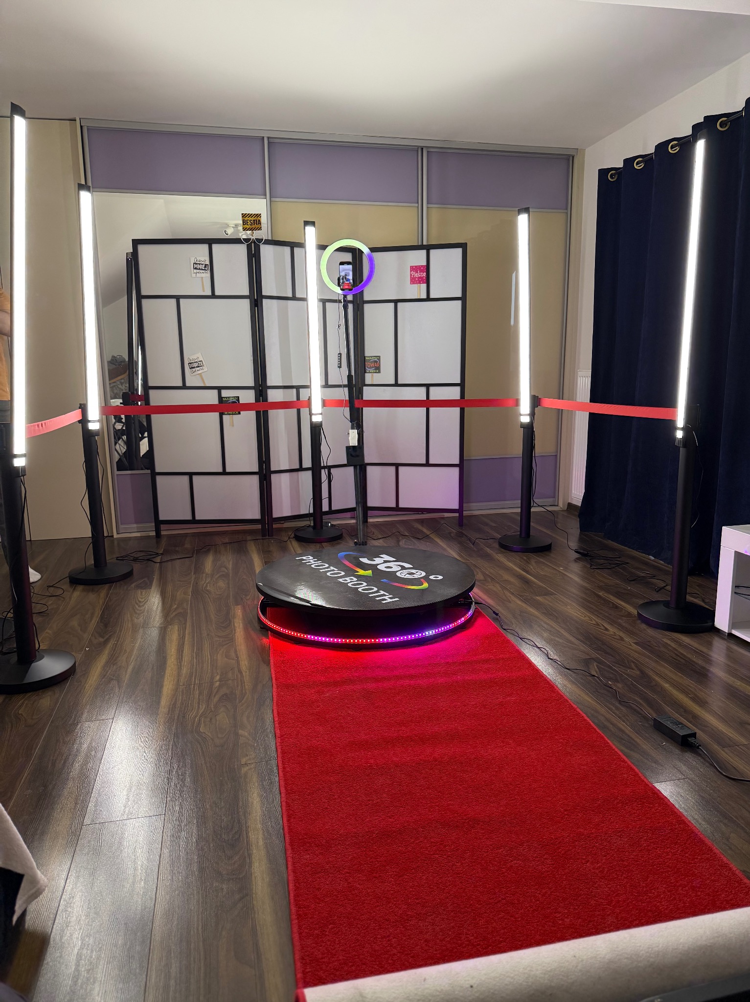 Oświetlona platforma obrotowa 360 stopni z czerwonym dywanem i barierkami oddzielającymi, otoczona lampami pierścieniowymi i tłem z dekoracjami.