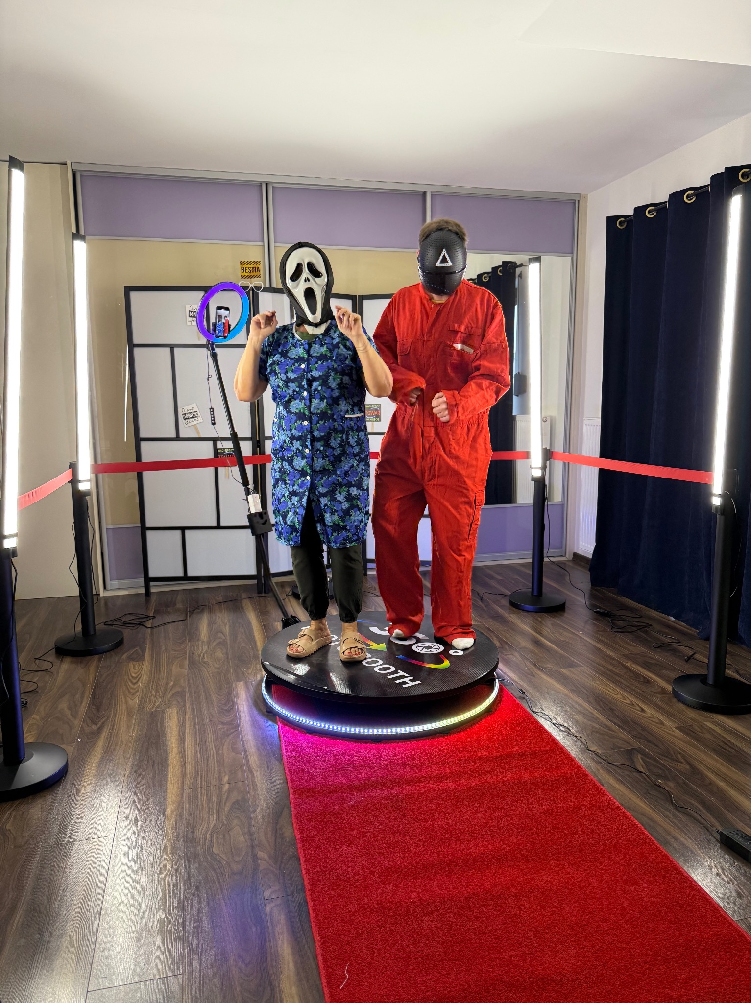 Dwie osoby w maskach, Scream i Squid Game, pozują na obrotowej platformie z oświetleniem LED i czerwonym dywanem, otoczone lampami studyjnymi.