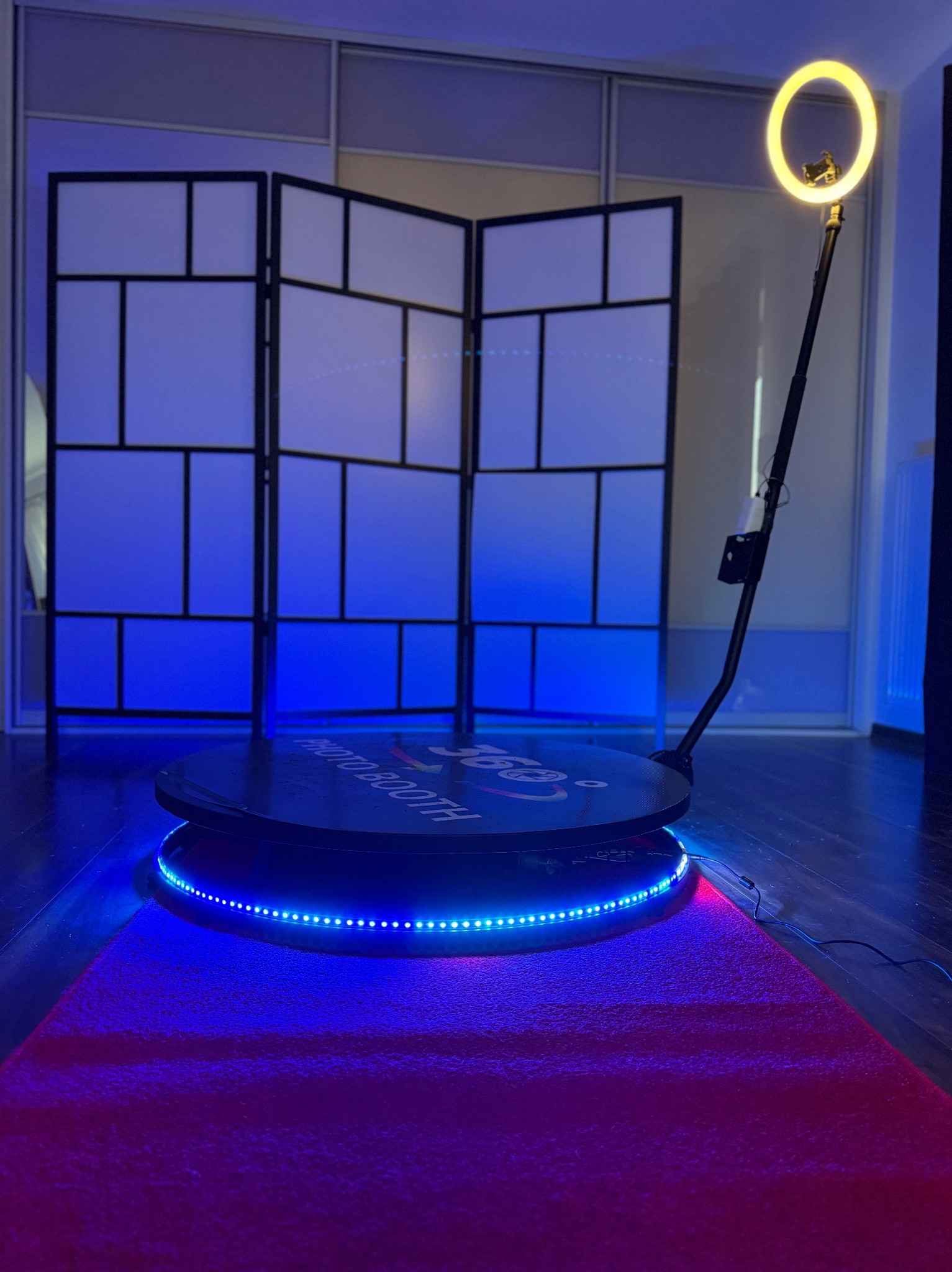 Oświetlona platforma 360 stopni z napisem PHOTO BOOTH, otoczona niebieskimi diodami LED, na różowym dywanie, z pierścieniową lampą i parawanem w tle.