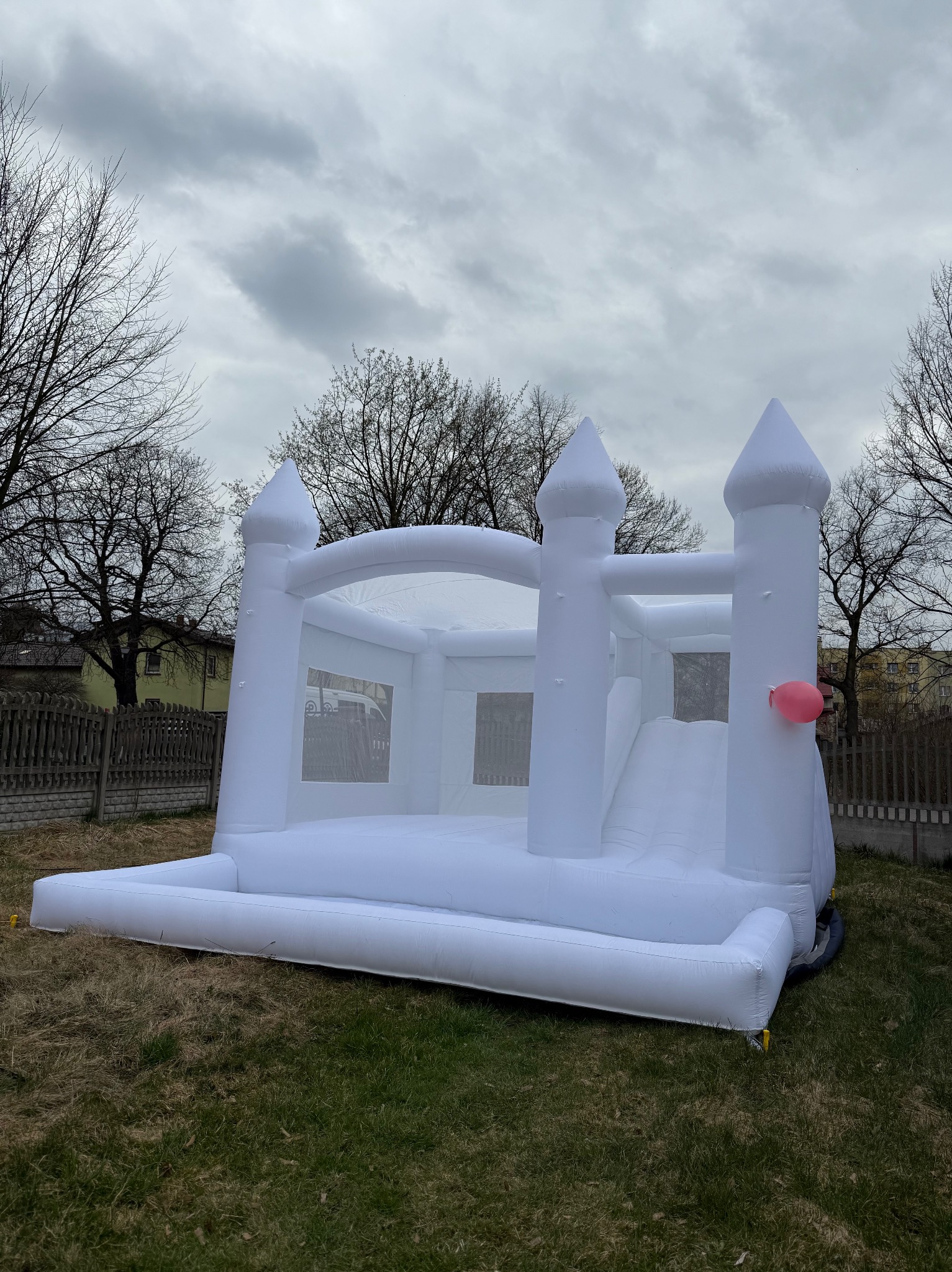 Biały dmuchany zamek z wieżami i zjeżdżalnią, ozdobiony różowym balonem, ustawiony na trawie pod pochmurnym niebem.