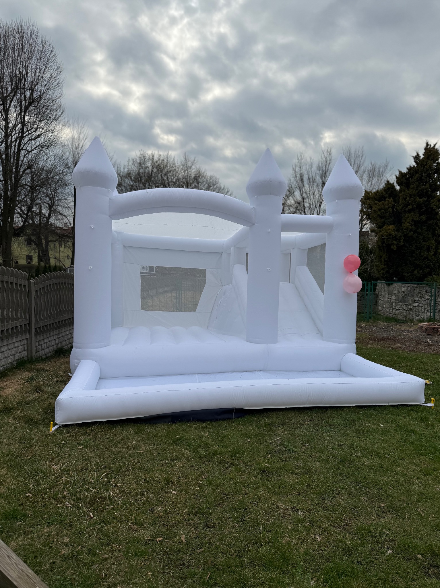 Biały dmuchany zamek z dwiema różowymi balonami na trawie w Jaworznie, ustawiony na tle pochmurnego nieba.