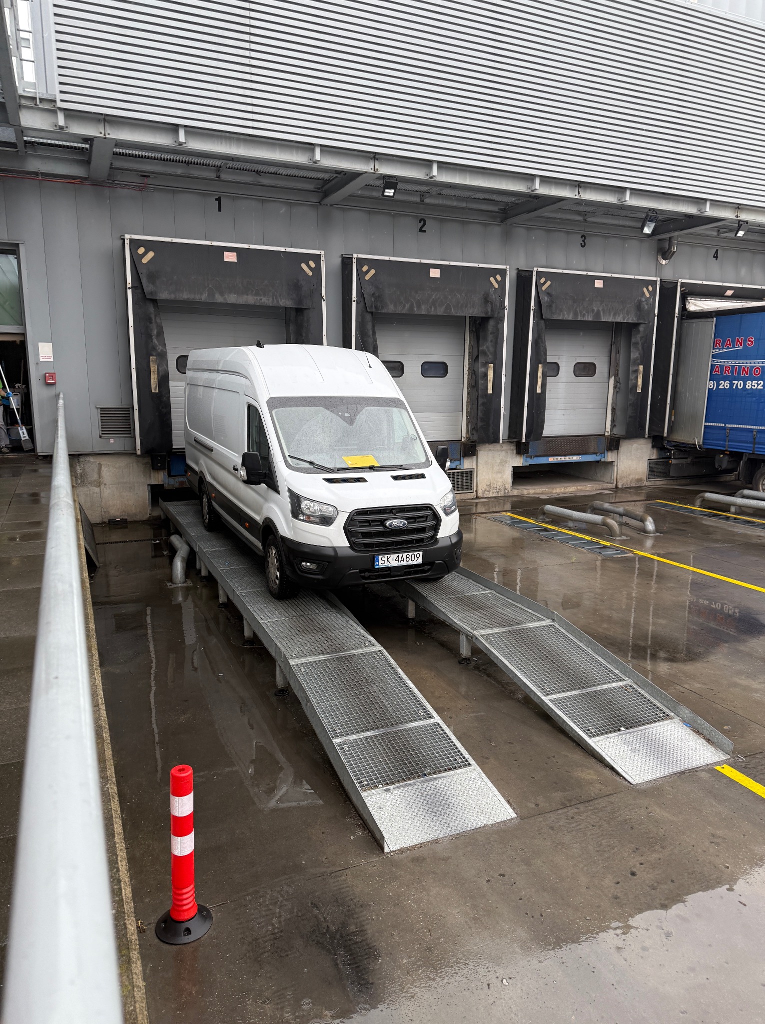 Biały bus Ford Transit na rampie załadunkowej, mokry plac, magazyn z bramami. Żółta kartka za szybą. Widoczne numery stanowisk.