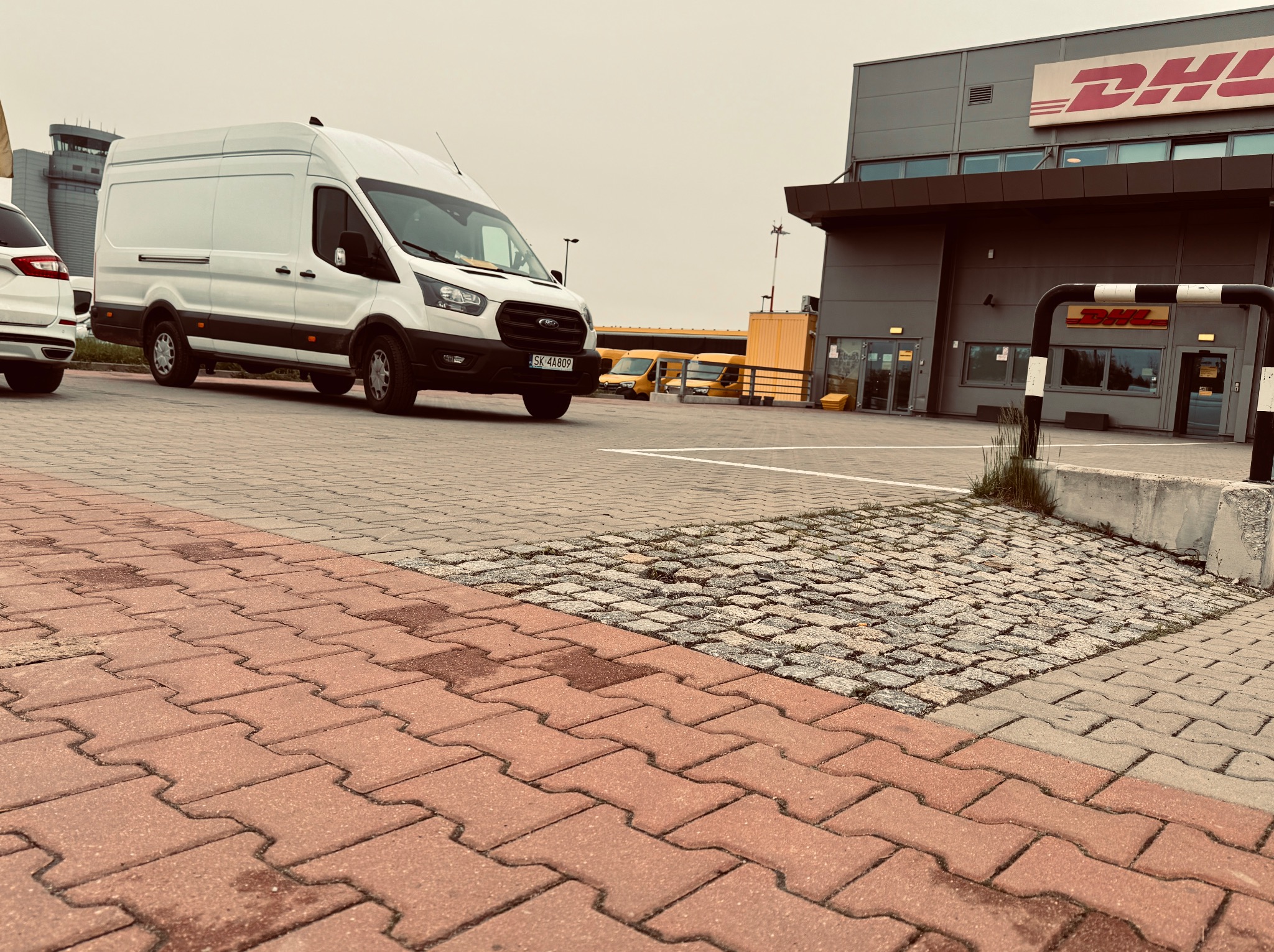 Biały bus dostawczy na tle budynku DHL, z żółtymi busami w tle. Widoczny fragment parkingu z kostki brukowej i fragment wieży kontroli lotów w oddali.