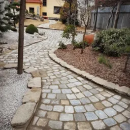 Kręta ścieżka z kostki brukowej w ogrodzie z dekoracyjnym żwirem, niskimi drzewami i krzewami, prowadząca do domu.