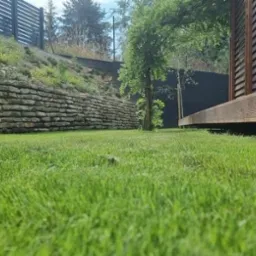 Nisko położona perspektywa na świeżo skoszoną, intensywnie zieloną trawę, z kamiennym murem oporowym na zboczu w tle i fragmentem drewnianej konstrukcji z przesłonami po prawej stronie.