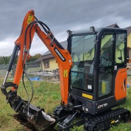 Od projektu do efektu - Pomarańczowa mini koparka Doosan DX19 na gąsienicach, stojąca na trawiastym terenie z widocznym domem w tle, pochmurne niebo.