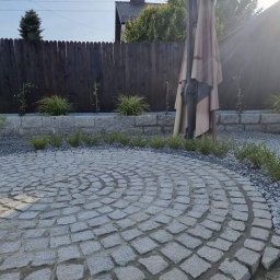 Od projektu do efektu - Okrągły taras z szarej kostki granitowej ułożonej w spiralny wzór, otoczony rabatami z niskimi roślinami i jasnym żwirem. W tle drewniany płot i złożony parasol.