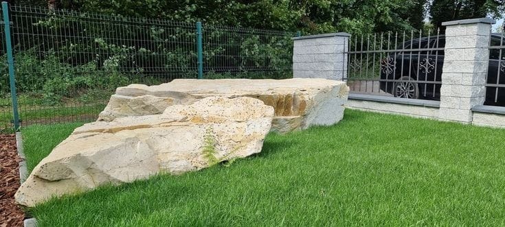 Duży, nieregularny blok piaskowca na trawniku w Ślemieniu, z widocznymi warstwami i porowatą strukturą, na tle ogrodzenia panelowego i fragmentu samochodu.