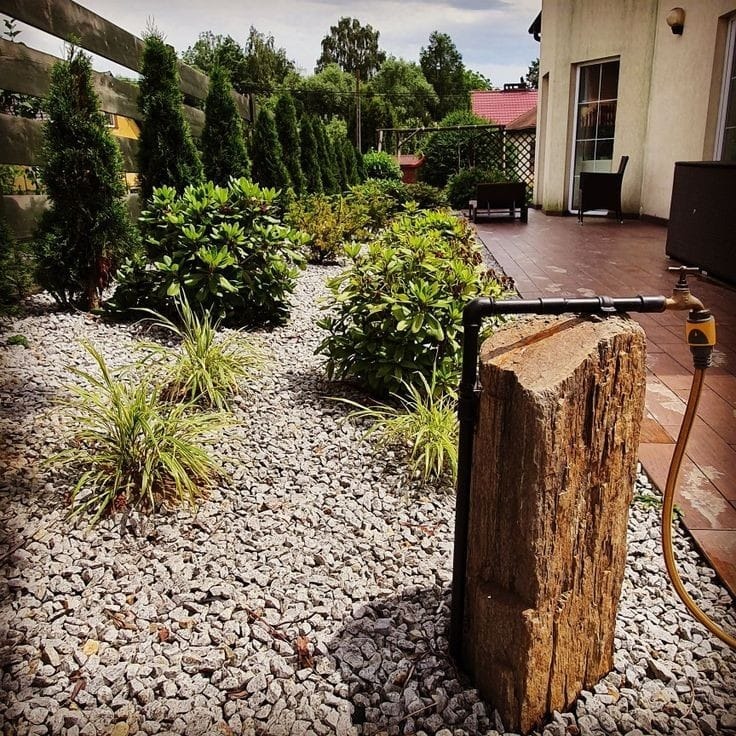 Ogród z dekoracyjnym ujęciem wody z kamienia, otoczony żwirem, z roślinami ozdobnymi i tujami w tle, drewniany płot po lewej stronie, taras z meblami ogrodowymi po prawej.