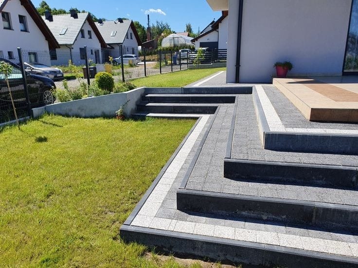 Schody i ścieżka z kostki brukowej w odcieniach szarości, z białymi akcentami, prowadzące do domu z trawnikiem i zielenią w tle.