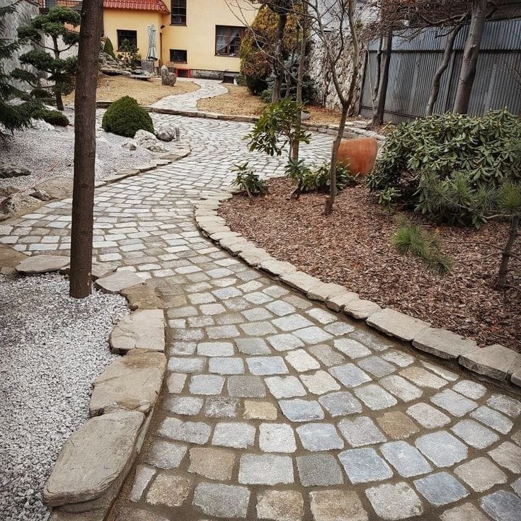 Kręta ścieżka z kostki brukowej w ogrodzie z dekoracyjnym żwirem, niskimi drzewami i krzewami, prowadząca do domu.