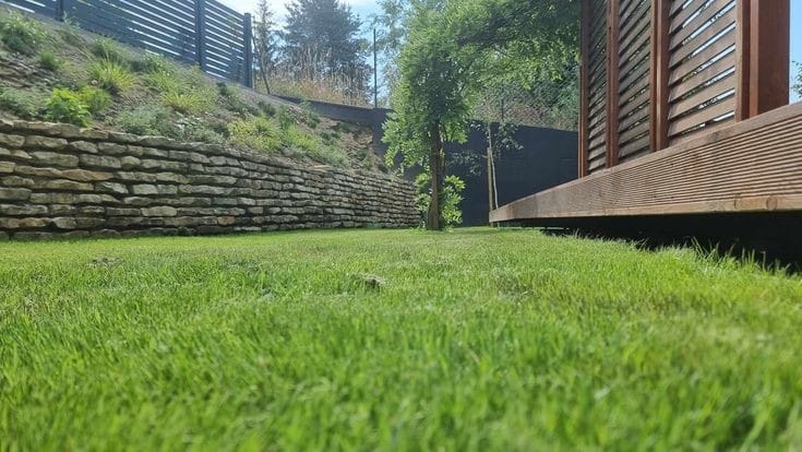 Nisko położona perspektywa na świeżo skoszoną, intensywnie zieloną trawę, z kamiennym murem oporowym na zboczu w tle i fragmentem drewnianej konstrukcji z przesłonami po prawej stronie.