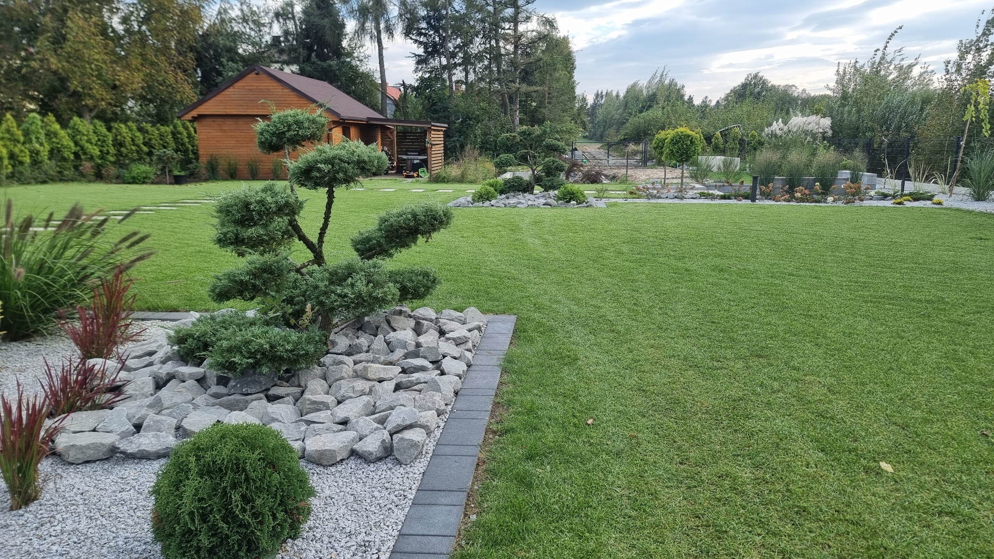 Zadbany ogród z równo skoszoną trawą, ozdobną roślinnością na skalniaku z kamieni i drewnianą altaną w tle.