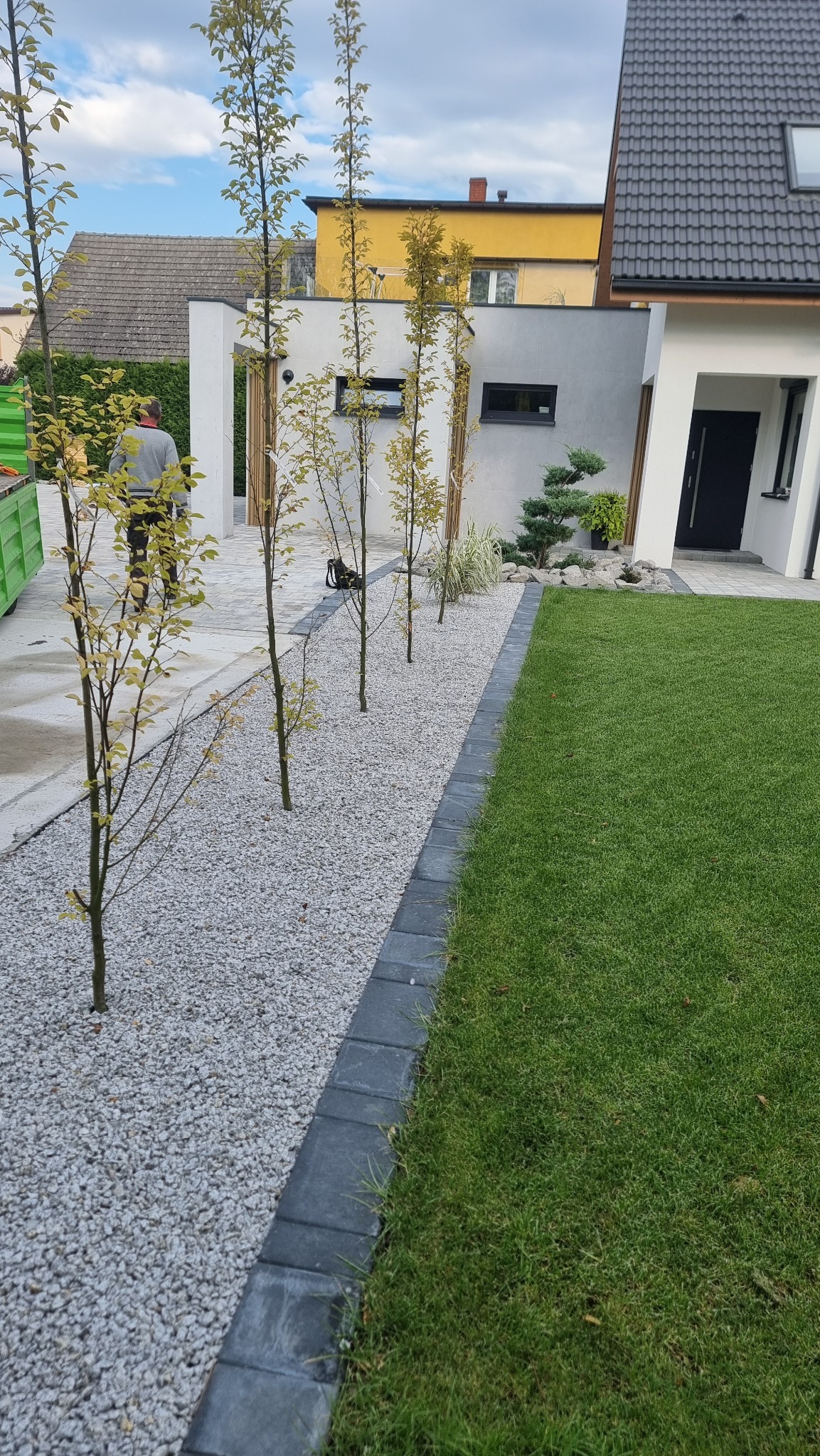 Nowoczesny ogród z równo przystrzyżonym trawnikiem, szarym żwirem oddzielonym obrzeżem z kostki brukowej, młodymi drzewkami i elementami małej architektury w tle.