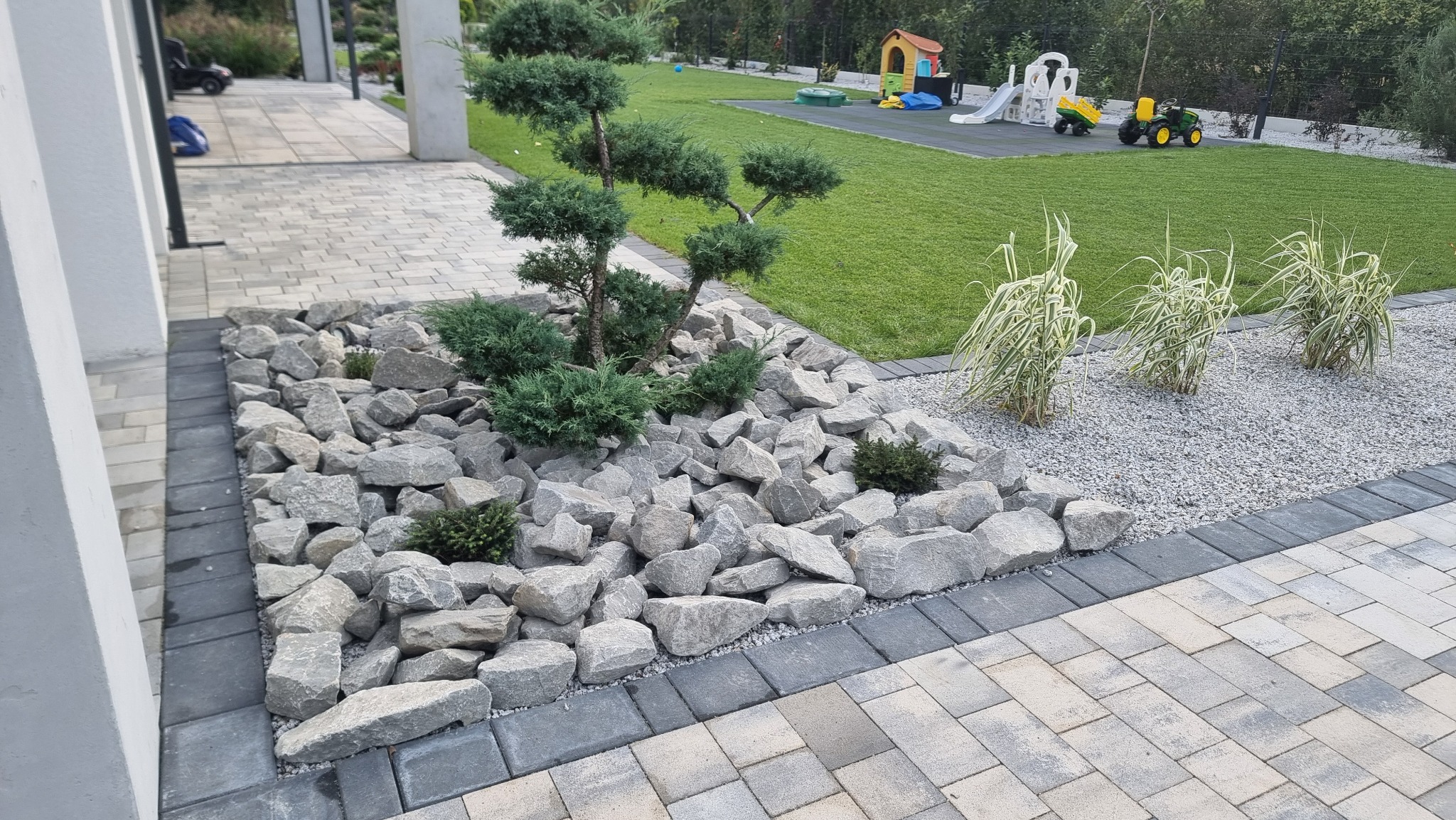 Ogród z ozdobnym drzewkiem bonsai otoczonym kamieniami, trawnikiem i placem zabaw w tle, obrzeża z kostki brukowej.