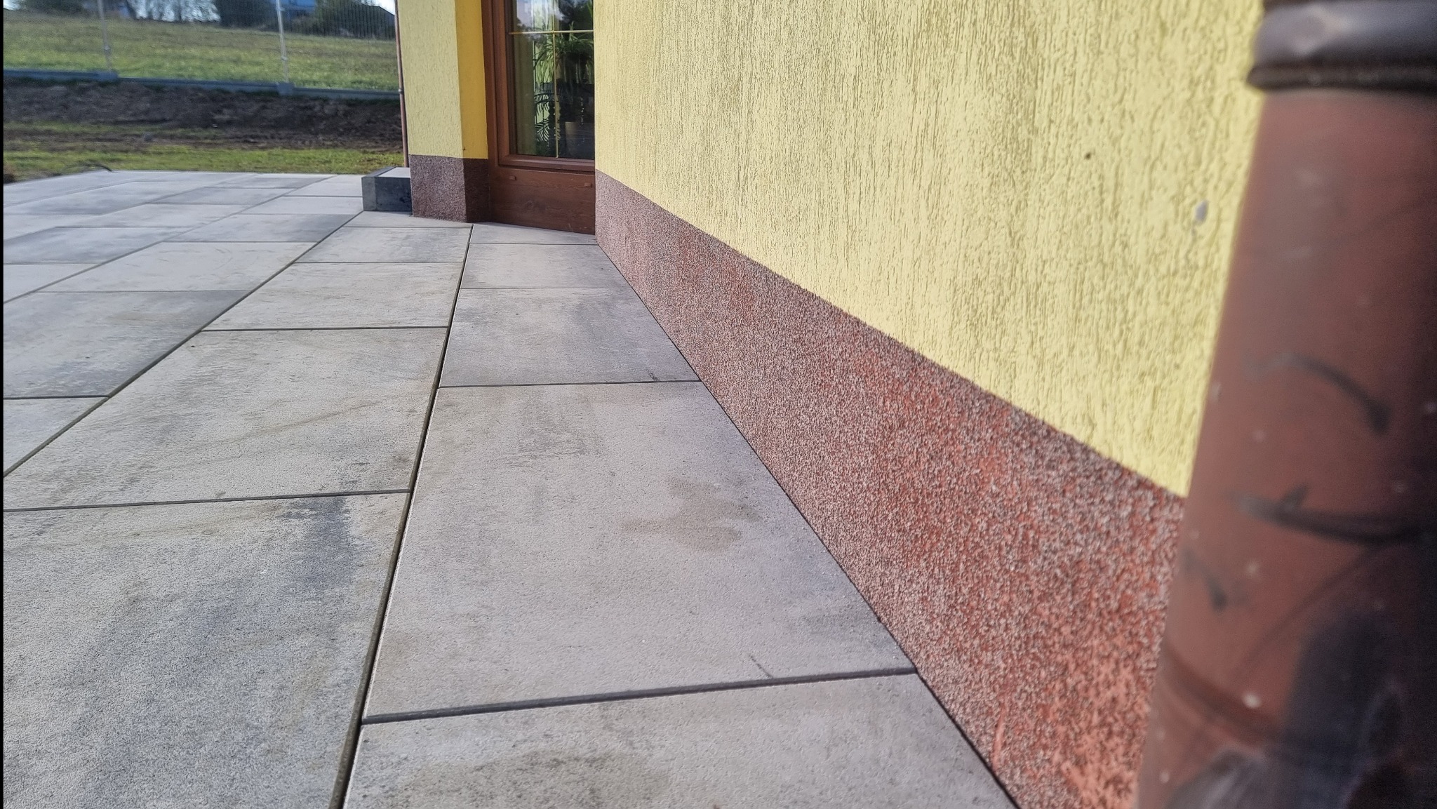 Szare płyty chodnikowe ułożone przy elewacji budynku z żółtym tynkiem i brązowym cokołem, widoczna rynna spustowa.