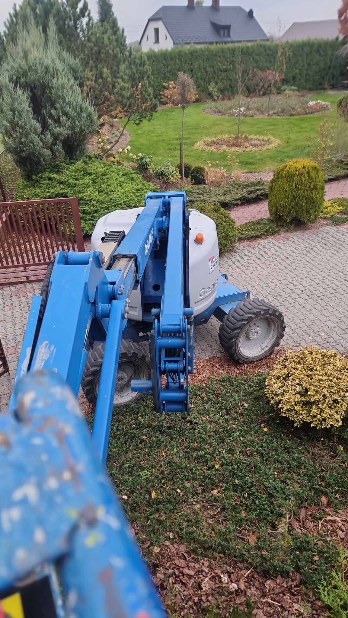 Widok z dołu na podnośnik koszowy Genie w ogrodzie, z widocznymi elementami krajobrazu i fragmentem domu w tle.
