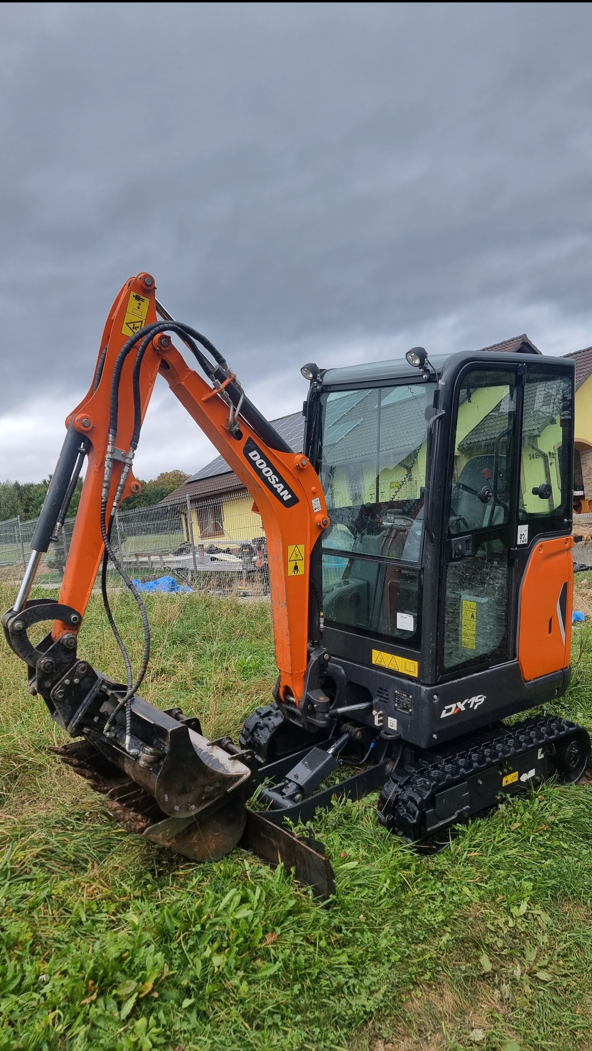 Pomarańczowa mini koparka Doosan DX19 na gąsienicach, stojąca na trawiastym terenie z widocznym domem w tle, pochmurne niebo.