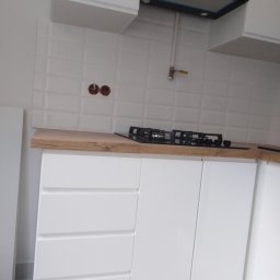 Aleks - Białe, minimalistyczne szafki kuchenne z drewnianym blatem i płytą gazową w mieszkaniu w Warszawie. Nad nimi białe, wypukłe płytki i czarny okap.