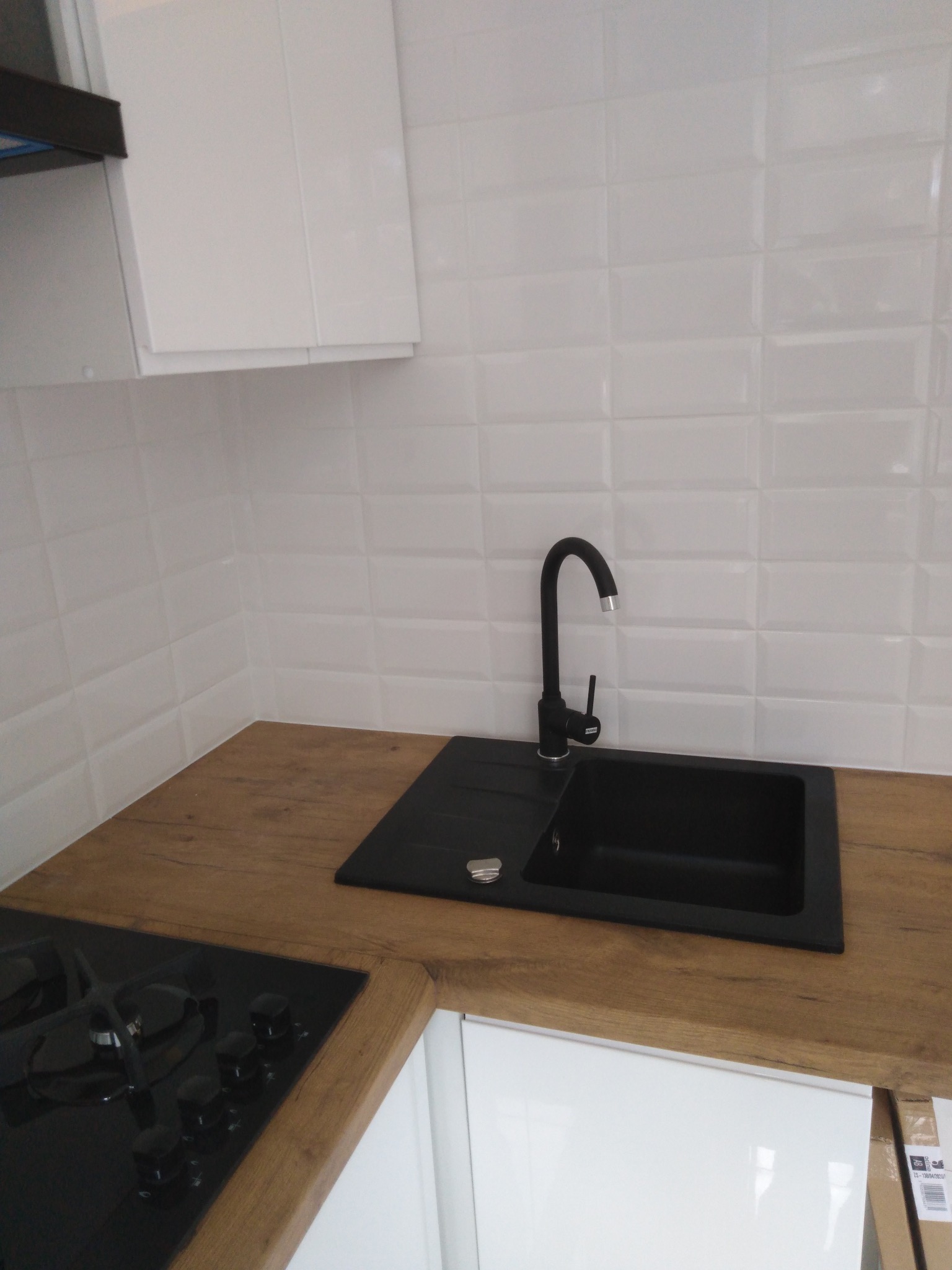 Nowoczesna kuchnia: białe szafki, drewniany blat, czarny zlew i bateria na tle białych, błyszczących płytek w stylu cegiełek. Widoczna płyta grzewcza.