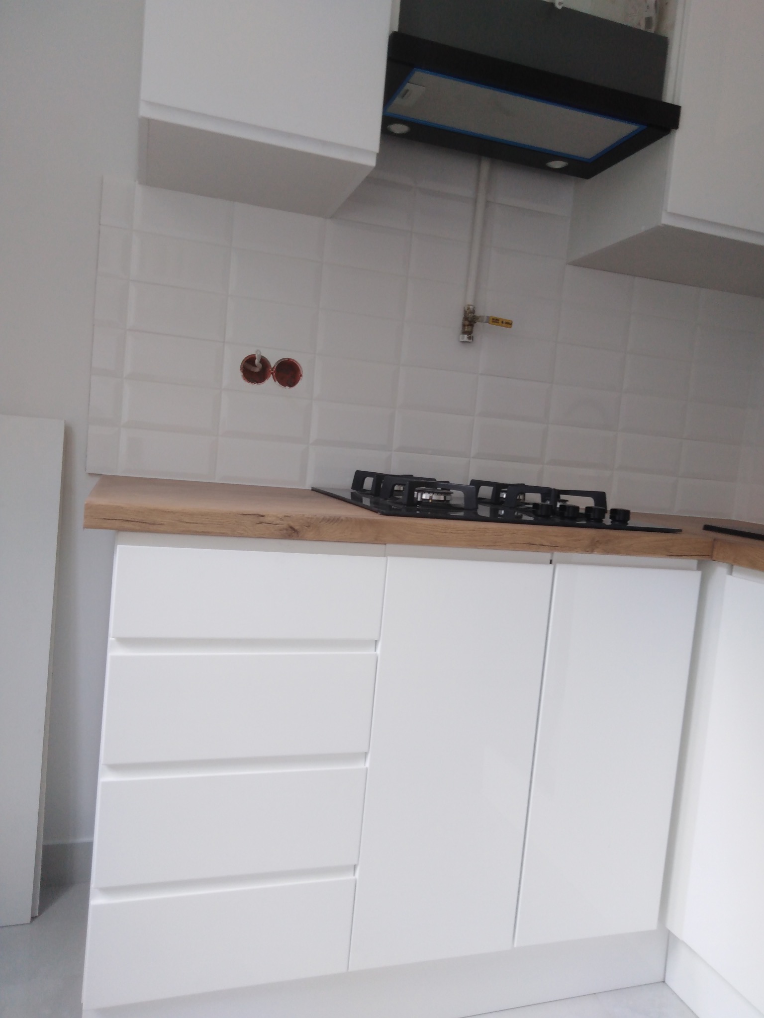 Białe, minimalistyczne szafki kuchenne z drewnianym blatem i płytą gazową w mieszkaniu w Warszawie. Nad nimi białe, wypukłe płytki i czarny okap.