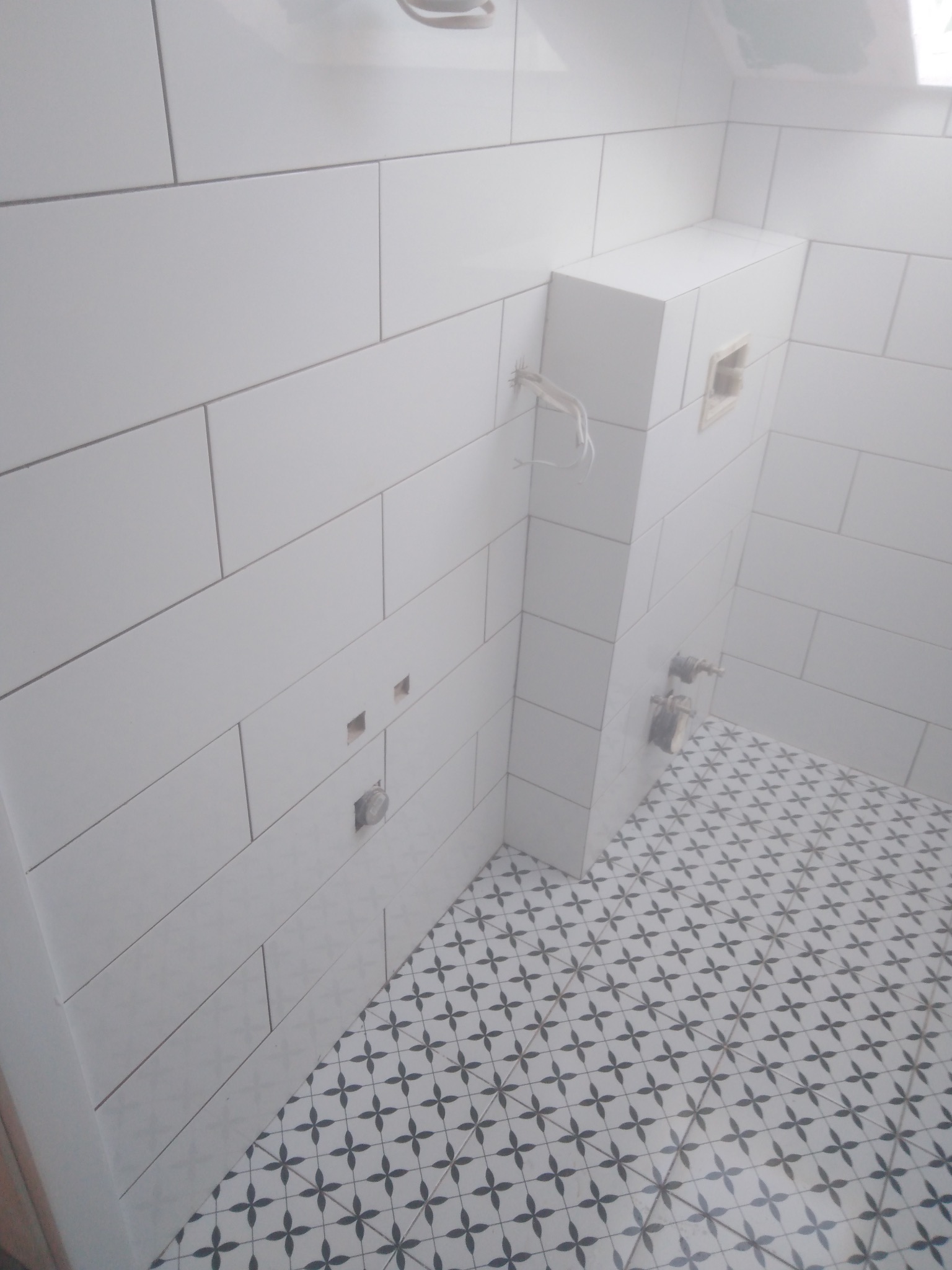 Białe, prostokątne płytki ścienne w łazience, z widocznymi instalacjami i wzorzystą podłogą w kwiatowy deseń. Częściowo wykończona zabudowa WC.