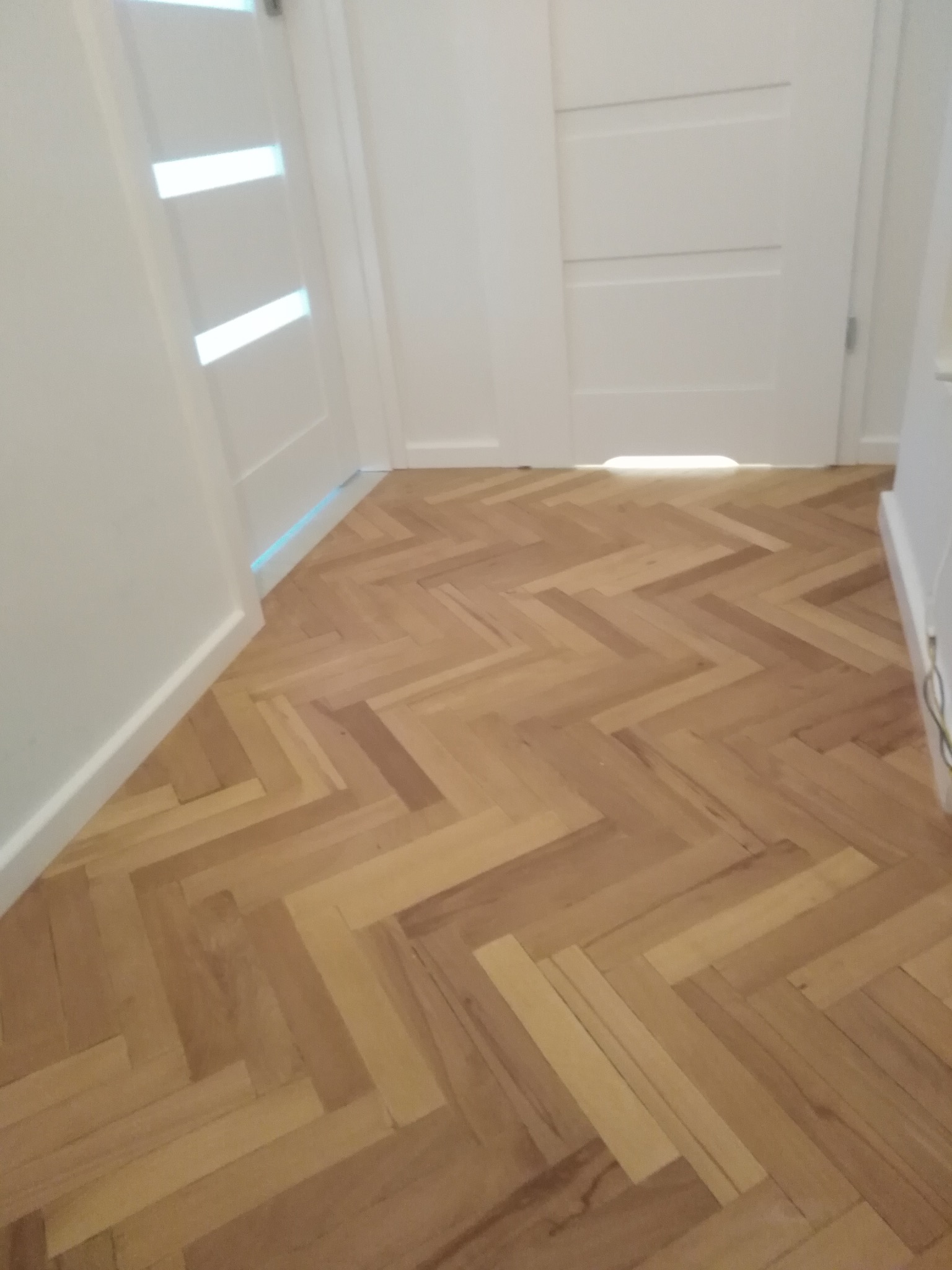 Parkiet ułożony w jodełkę, jasne drewno, widoczne drzwi w tle. Naturalne oświetlenie podkreśla wzór podłogi. Detal wykończenia wnętrza.