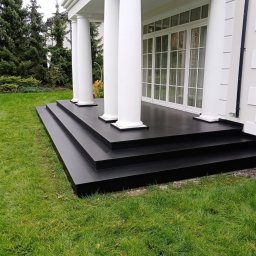 Volodymyr KUZ - Nowoczesny taras z czarnych płyt, z trzema stopniami, prowadzący do domu z białymi kolumnami i dużymi oknami, otoczony zielonym trawnikiem i drzewami iglastymi.