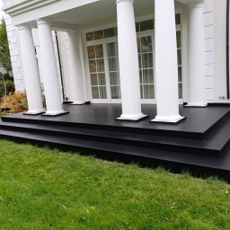 Volodymyr KUZ - Eleganckie wejście do domu z czarnymi schodami i tarasem, otoczone zielonym trawnikiem, białe kolumny podtrzymują zadaszenie.