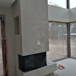 Volodymyr KUZ - Obudowa kominka z płyt kamiennych imitujących marmur, z widocznym otworem paleniska zabezpieczonym folią, w trakcie montażu w nowoczesnym salonie z dużymi oknami.
