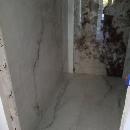 Volodymyr KUZ - Wnętrze łazienki w trakcie remontu, widoczne marmurowe płytki na ścianach i podłodze, surowy narożnik ściany z widocznym murem, odbicie sylwetki w błyszczącej powierzchni płytek.