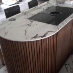 Volodymyr KUZ - Wyspa kuchenna z blatem z białego marmuru z ciemnymi żyłami, wbudowaną płytą grzewczą i obudową z pionowych drewnianych listew, na podłodze z marmuru.