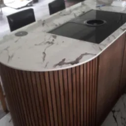 Wyspa kuchenna z blatem z białego marmuru z ciemnymi żyłami, wbudowaną płytą grzewczą i obudową z pionowych drewnianych listew, na podłodze z marmuru.