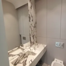 Nowoczesna łazienka z marmurową umywalką, blatem i podłogą w odcieniach bieli i szarości; minimalistyczne szafki i toaleta w tle.