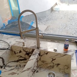 Volodymyr KUZ - Dwu-komorowy zlew z marmurowym wzorem i nowoczesną baterią z elastyczną wylewką, widok na okno z gruzem budowlanym na zewnątrz, zabezpieczone folią malarską, butelka płynu do czyszczenia na blacie.