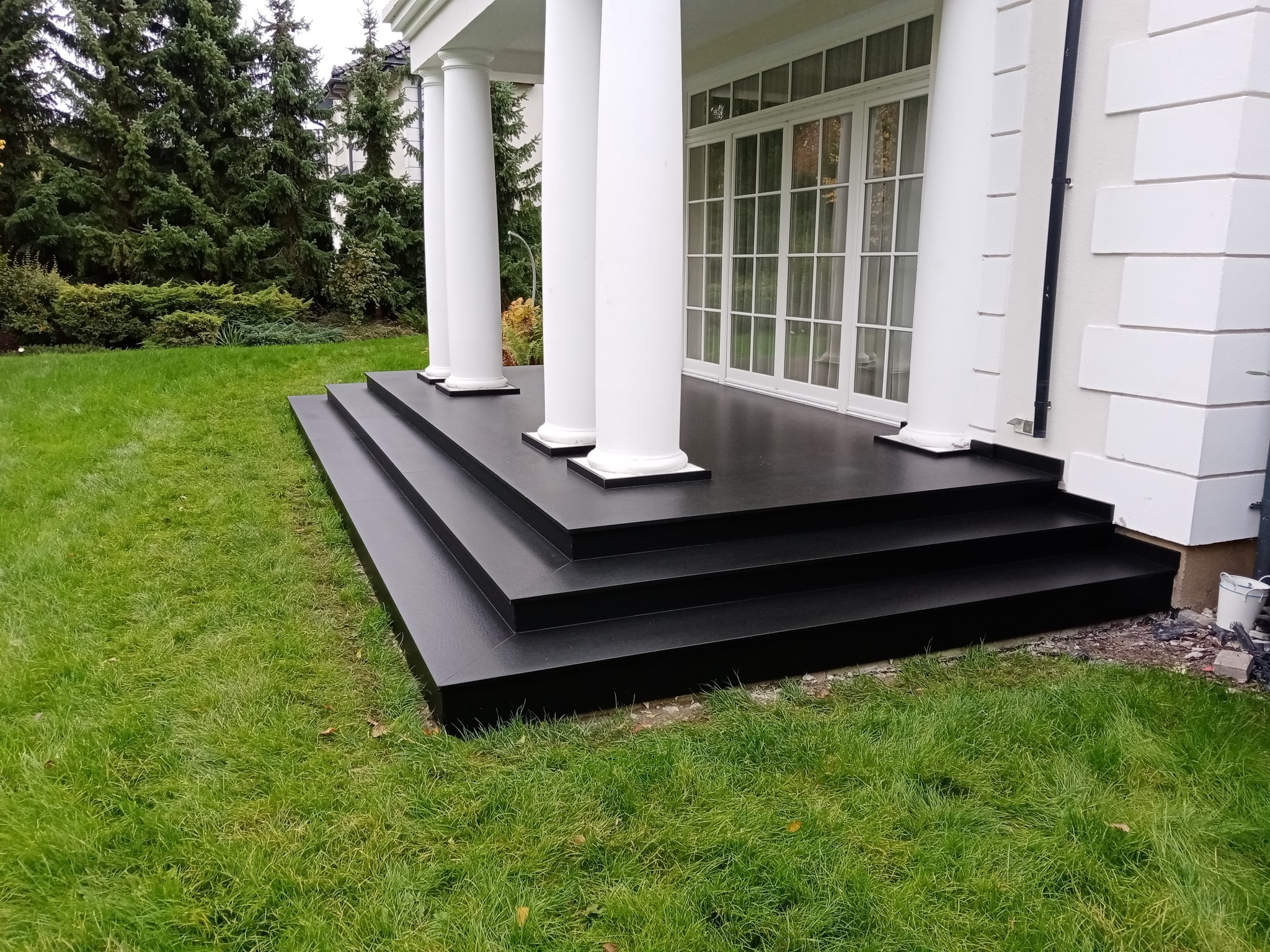 Nowoczesny taras z czarnych płyt, z trzema stopniami, prowadzący do domu z białymi kolumnami i dużymi oknami, otoczony zielonym trawnikiem i drzewami iglastymi.