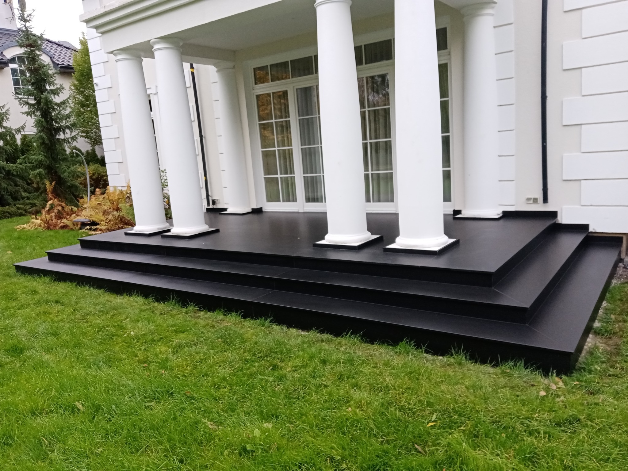 Eleganckie wejście do domu z czarnymi schodami i tarasem, otoczone zielonym trawnikiem, białe kolumny podtrzymują zadaszenie.