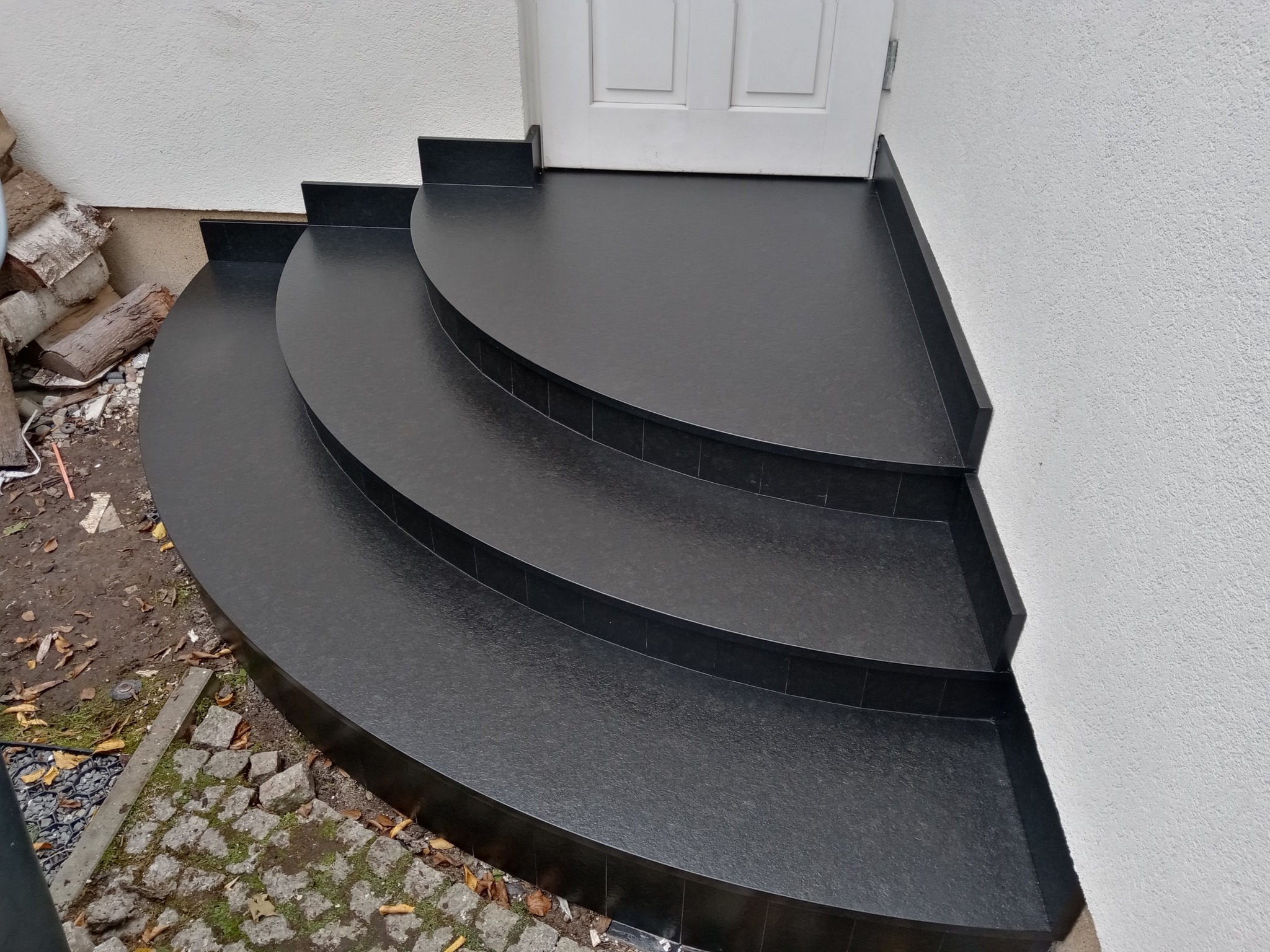 Trzy półokrągłe stopnie z czarnego kamienia prowadzące do białych drzwi wejściowych, widok z boku, z boku stos drewna.