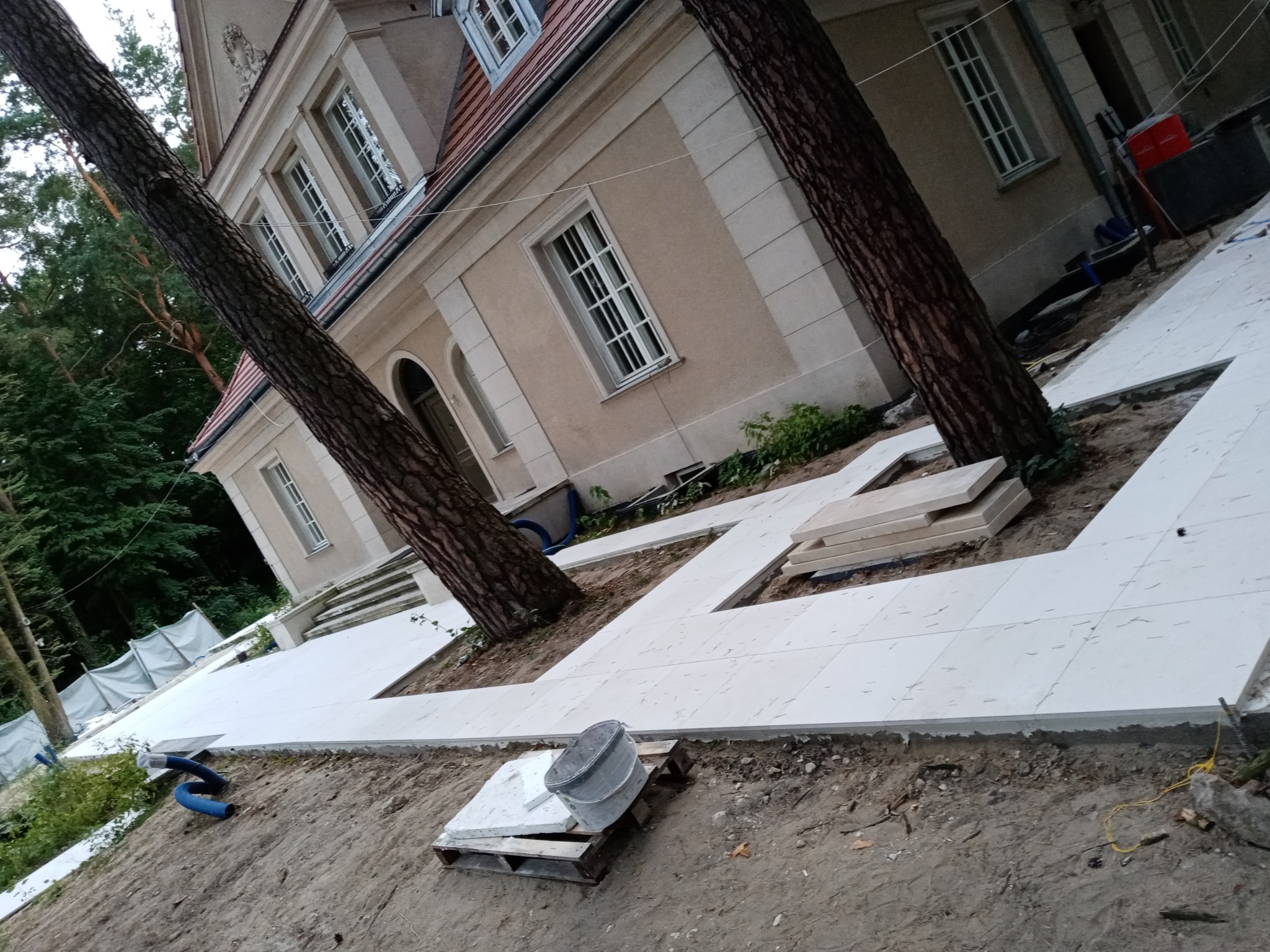 Układanie jasnych płyt tarasowych wokół drzew przy budynku, widoczny paleta z wiadrem i przygotowane elementy