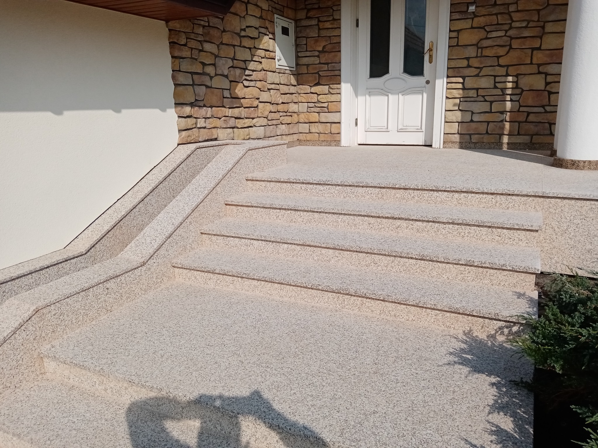 Granitowe schody zewnętrzne prowadzące do białych drzwi wejściowych, zlokalizowane w Pruszkowie, z elewacją wykończoną kamieniem naturalnym.