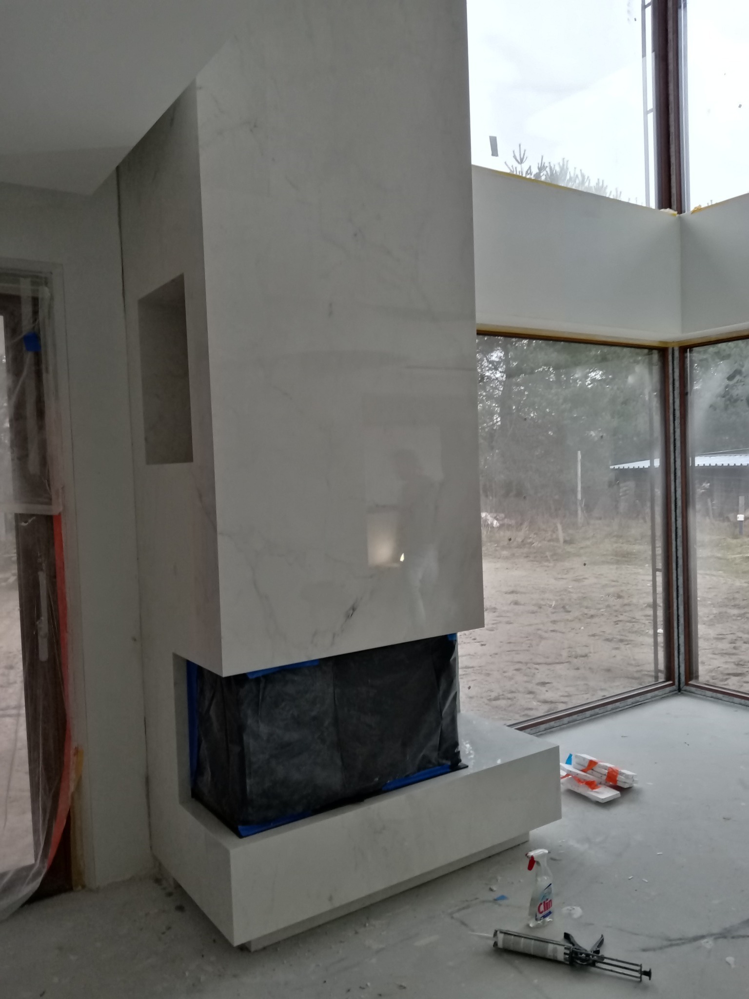 Obudowa kominka z płyt kamiennych imitujących marmur, z widocznym otworem paleniska zabezpieczonym folią, w trakcie montażu w nowoczesnym salonie z dużymi oknami.
