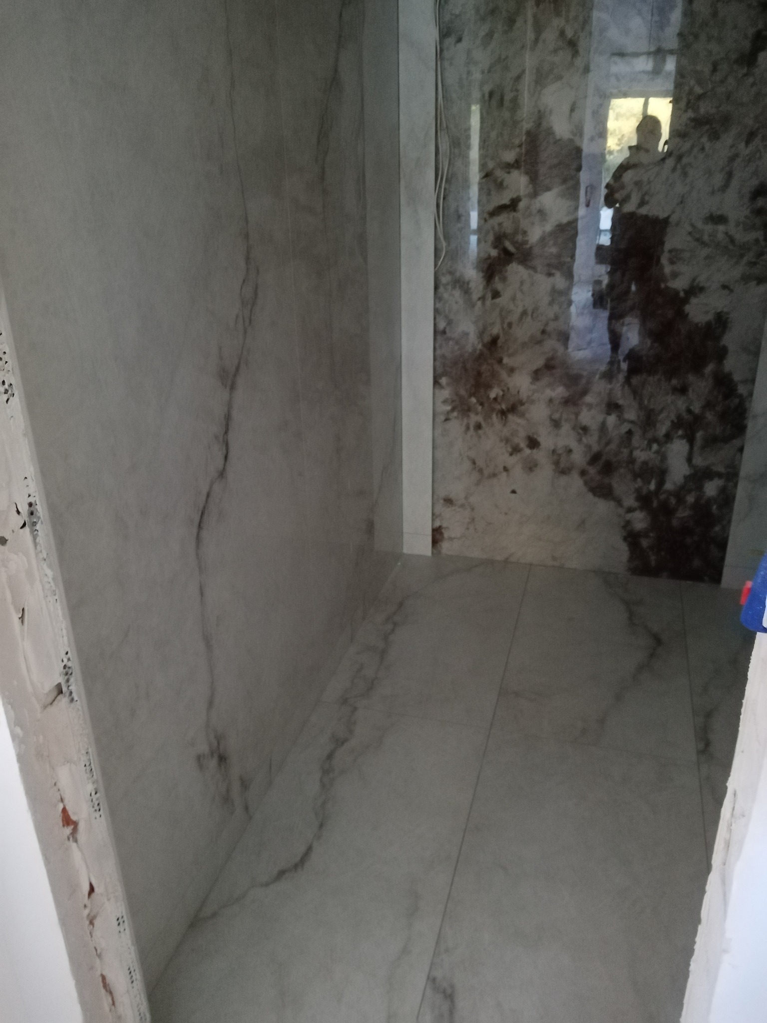 Wnętrze łazienki w trakcie remontu, widoczne marmurowe płytki na ścianach i podłodze, surowy narożnik ściany z widocznym murem, odbicie sylwetki w błyszczącej powierzchni płytek.