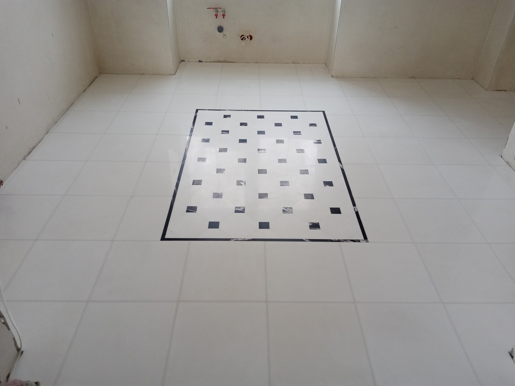 Biała podłoga z płytek z centralnie umieszczonym kwadratem z czarnymi akcentami, zabezpieczona folią ochronną w surowym wnętrzu łazienki przed montażem armatury.