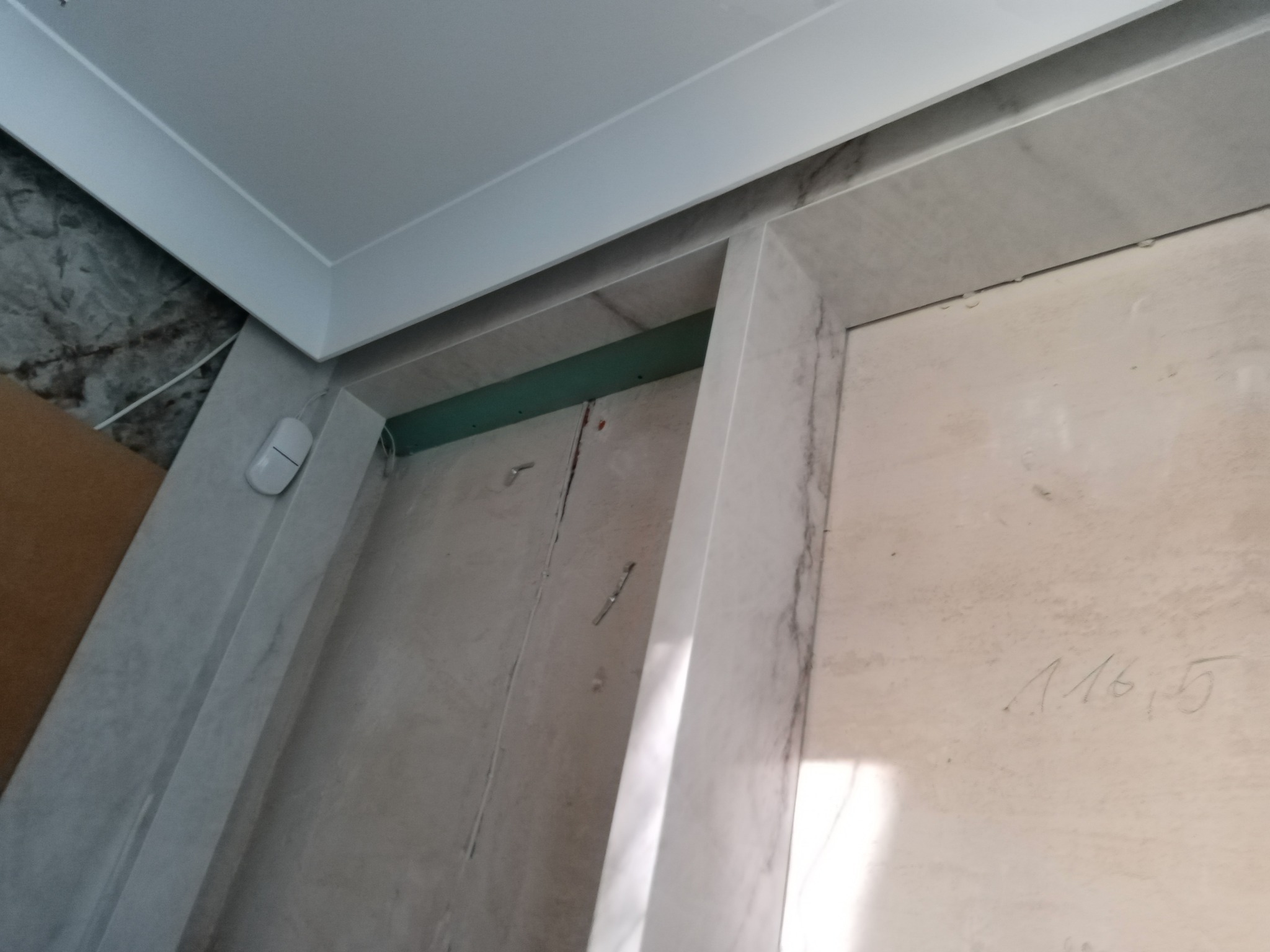 Wnętrze w trakcie prac wykończeniowych: widoczna ściana z płyt karton-gips, fragment sufitu z listwą, obłożone marmurem obramowanie drzwi oraz notatka ołówkiem na ścianie.