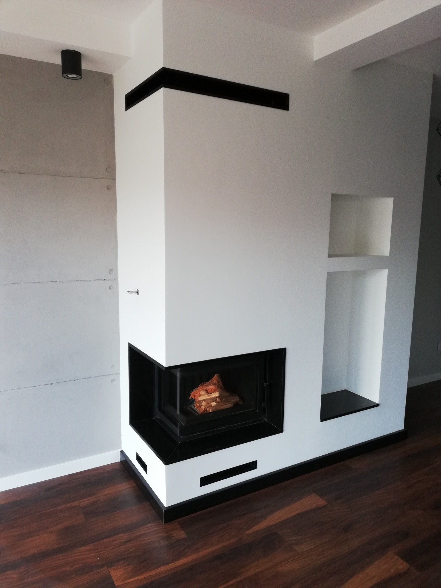 Nowoczesny kominek narożny z czarnymi akcentami, drewnem w palenisku i wnękami w ścianie obok, na tle betonowej ściany i ciemnej podłogi.