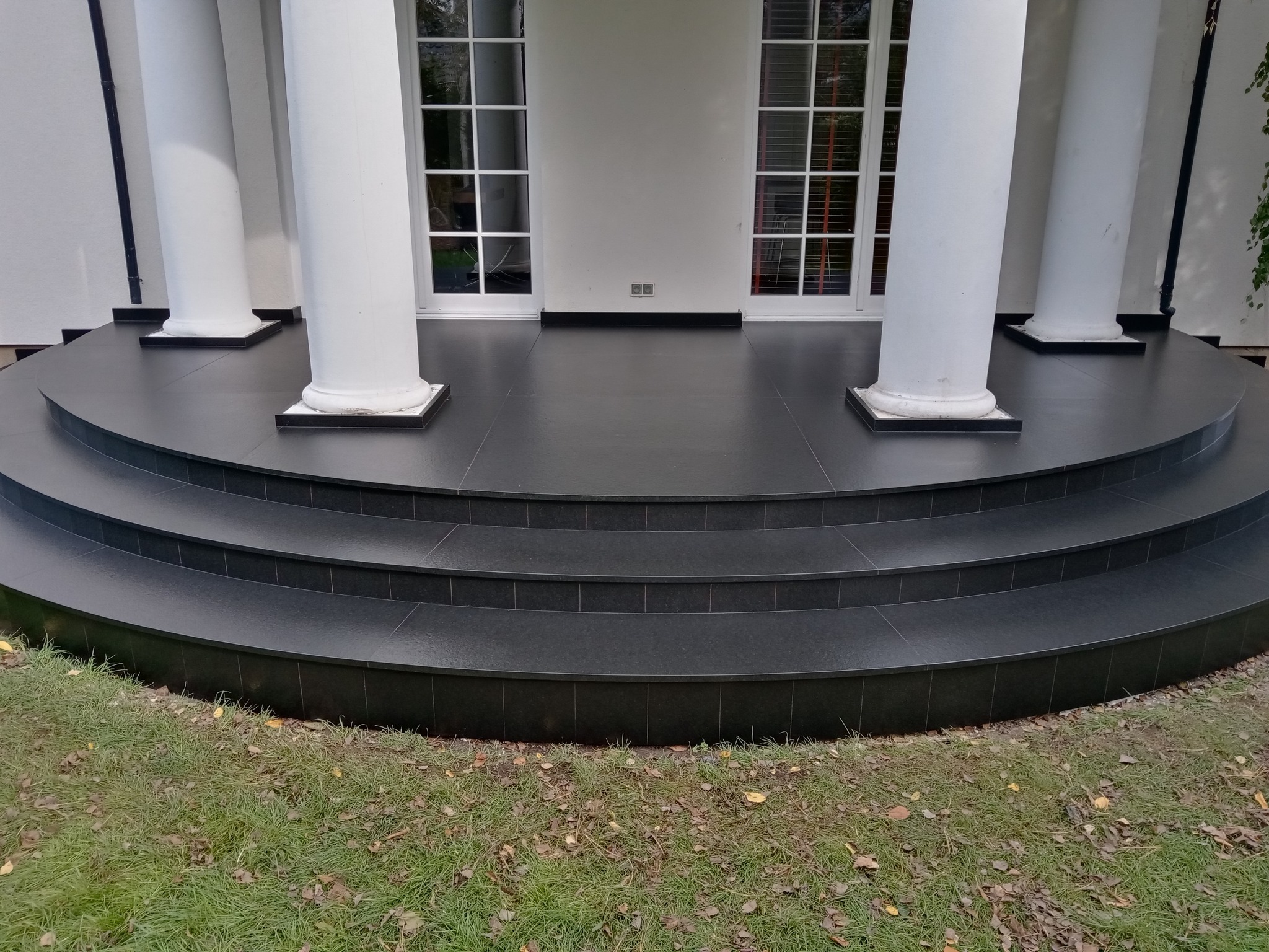 Elegancki taras z czarnych płytek kamiennych z trzema stopniami, otoczony białymi kolumnami i zielonym trawnikiem z opadłymi liśćmi.