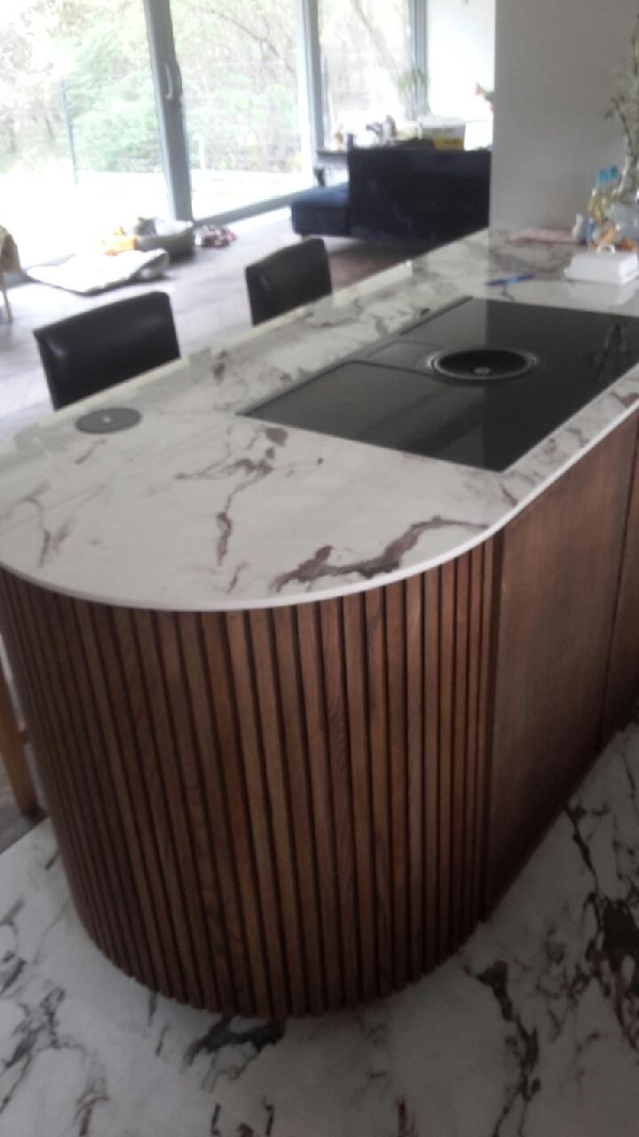 Wyspa kuchenna z blatem z białego marmuru z ciemnymi żyłami, wbudowaną płytą grzewczą i obudową z pionowych drewnianych listew, na podłodze z marmuru.